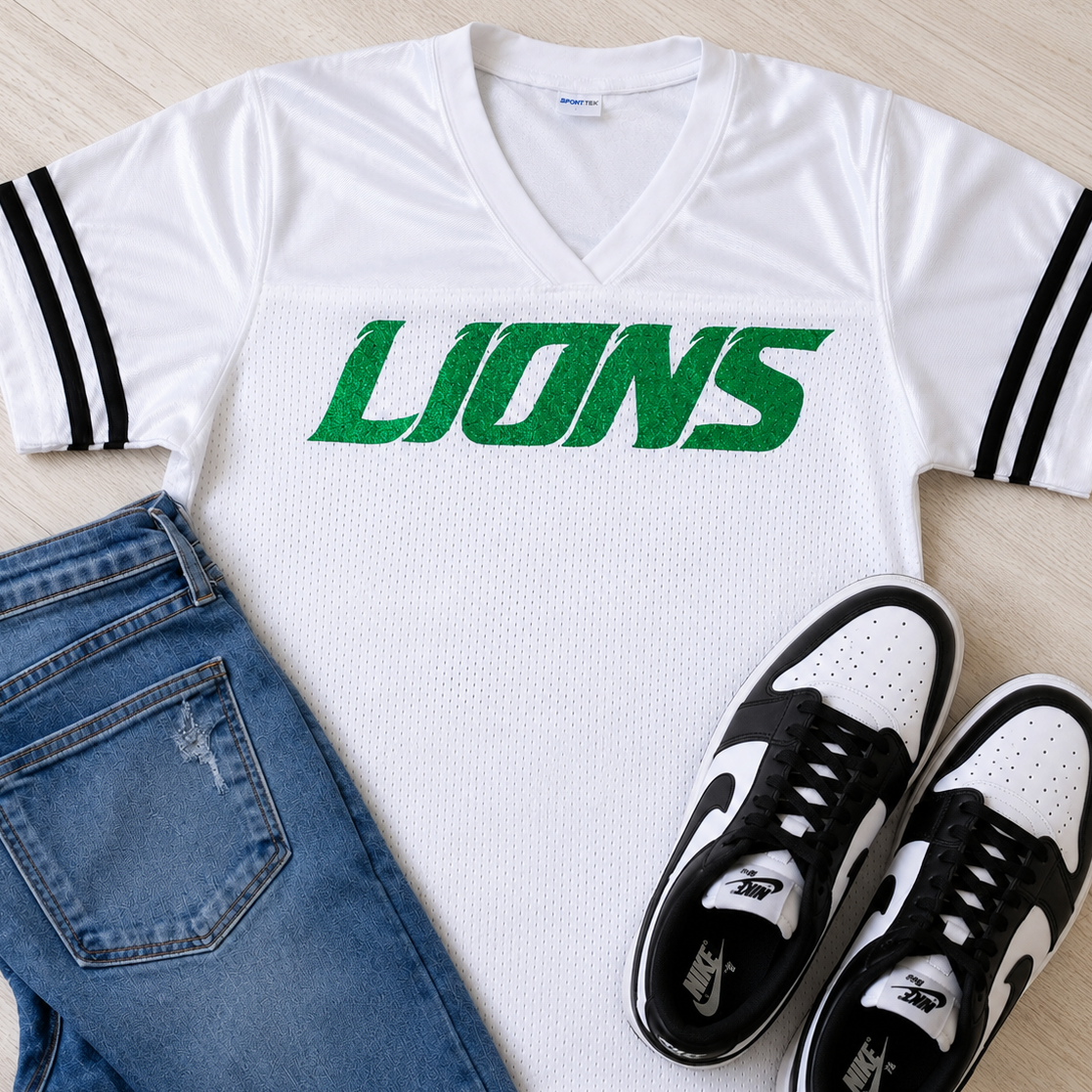 A white mesh V-neck sports jersey with black sleeve stripes and a green 'LIONS' graphic across the chest, styled in a flat lay with blue jeans and black-and-white sneakers on a light wood floor.