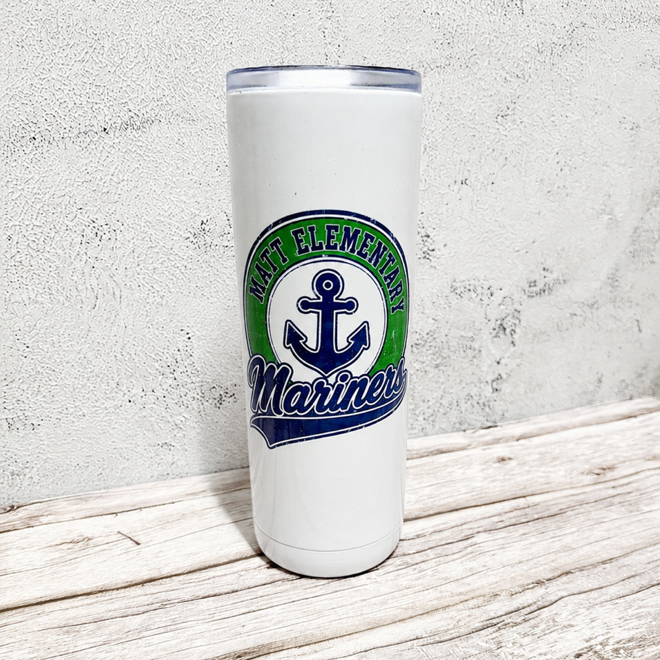A white insulated tumbler with a clear lid stands on a rustic light wood surface against a textured light gray wall. The tumbler features a circular 'North Elementary Mariners' graphic with a navy blue anchor and green and blue lettering.