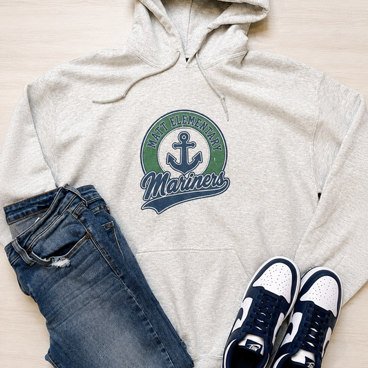 A flat-lay outfit arrangement featuring a light heather gray hoodie with a green and navy 'Fremont Mariners' graphic, paired with blue jeans and navy and white sneakers on a light wood background.