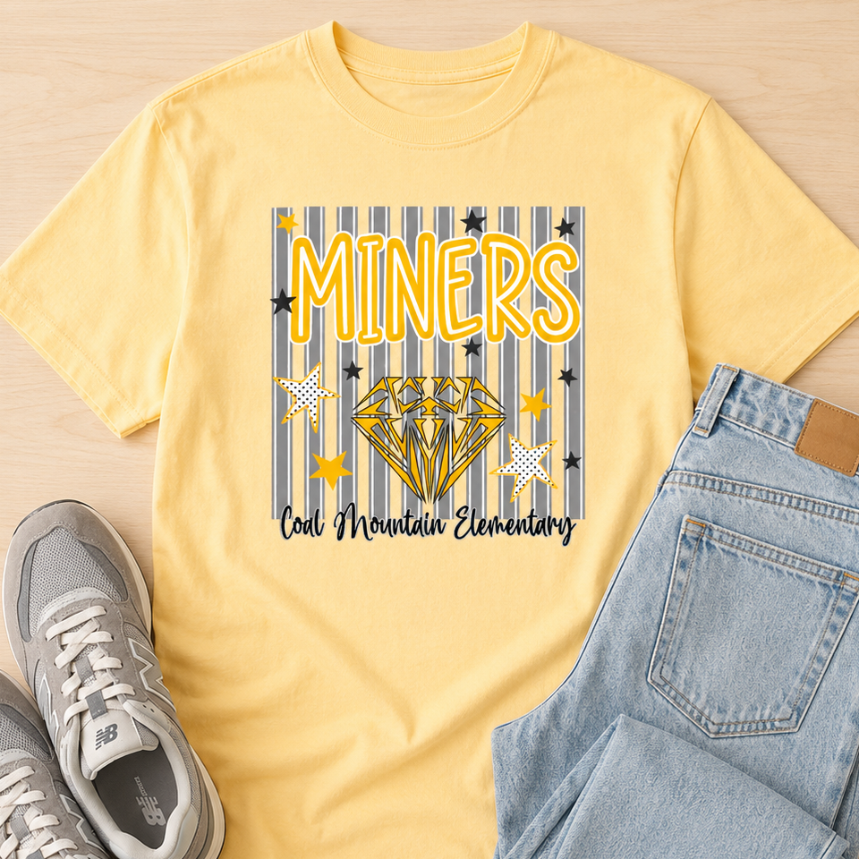 A yellow graphic t-shirt laid flat on a light wood background, styled with light blue jeans and gray sneakers. The t-shirt features a graphic with the word 'MINERS' in yellow, a gold diamond, and the text 'Coal Mountain Elementary' on a blue and yellow striped background.
