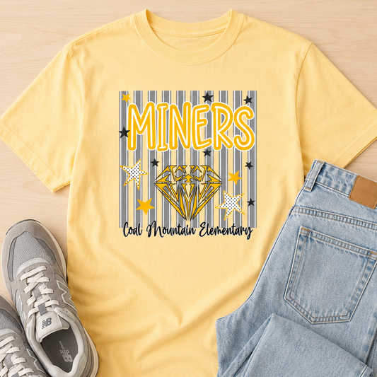 A yellow graphic t-shirt laid flat on a light wood background, styled with light blue jeans and gray sneakers. The t-shirt features a graphic with the word 'MINERS' in yellow, a gold diamond, and the text 'Coal Mountain Elementary' on a blue and yellow striped background.