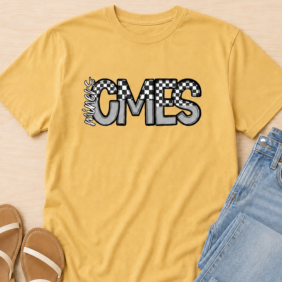 A yellow graphic t-shirt featuring the text 'INTERSTATE CMES' in a gray and black checkered font, styled in a flat lay with light blue jeans and tan sandals on a light wood background.