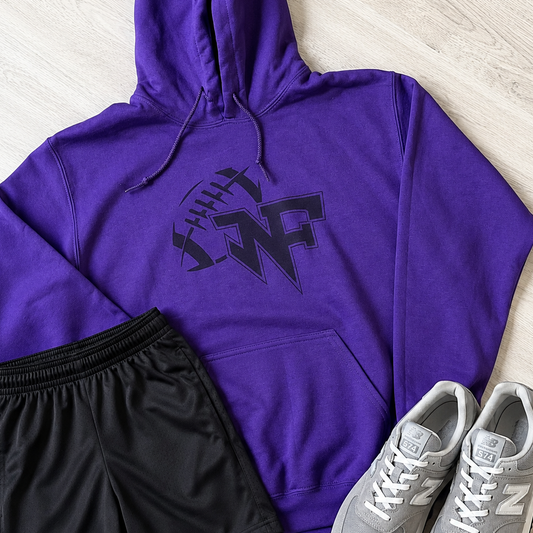 A purple pullover hoodie with a black graphic logo on the chest, laid flat on a light wood floor alongside black athletic shorts and gray New Balance sneakers.