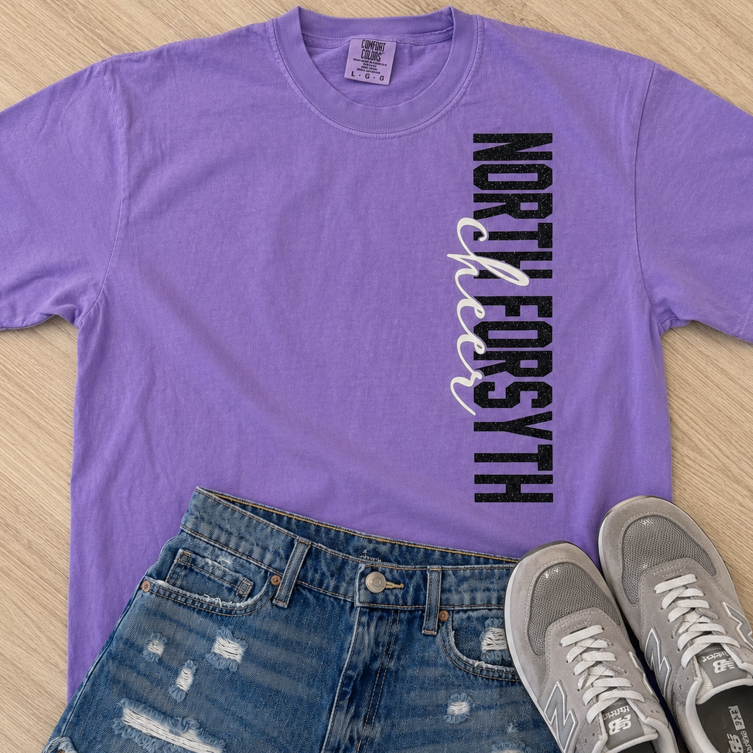 A purple short-sleeve graphic T-shirt laid flat on a light wood surface, paired with distressed blue denim shorts and gray New Balance sneakers. The shirt has vertical text reading 'NORTH FORSYTH' in black with a white and purple ribbon graphic.