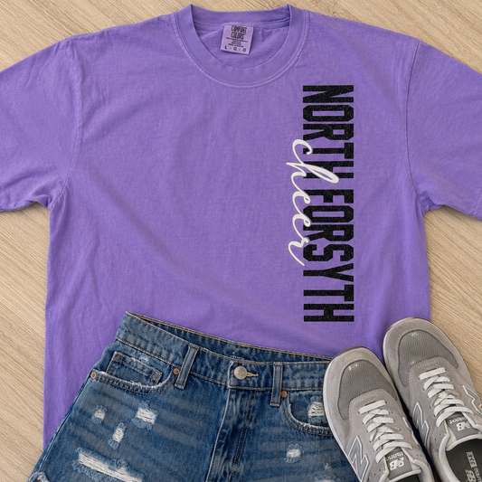 A purple short-sleeve graphic T-shirt laid flat on a light wood surface, paired with distressed blue denim shorts and gray New Balance sneakers. The shirt has vertical text reading 'NORTH FORSYTH' in black with a white and purple ribbon graphic.
