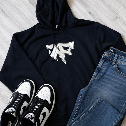 A styled flat-lay of a black hoodie with a white graphic logo, paired with blue jeans and black-and-white sneakers on a light wood floor.