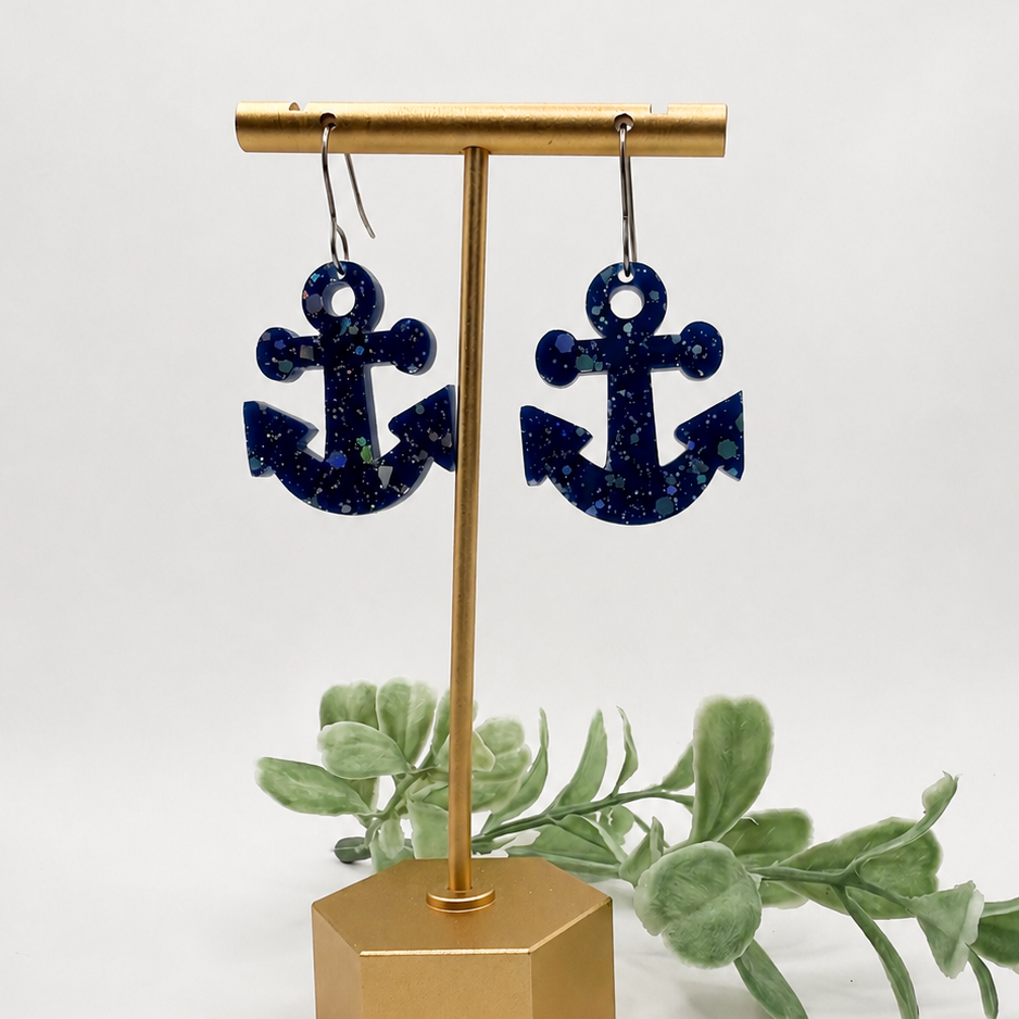 A pair of navy blue glitter anchor drop earrings displayed on a gold-tone stand with decorative green leaves against a white background.