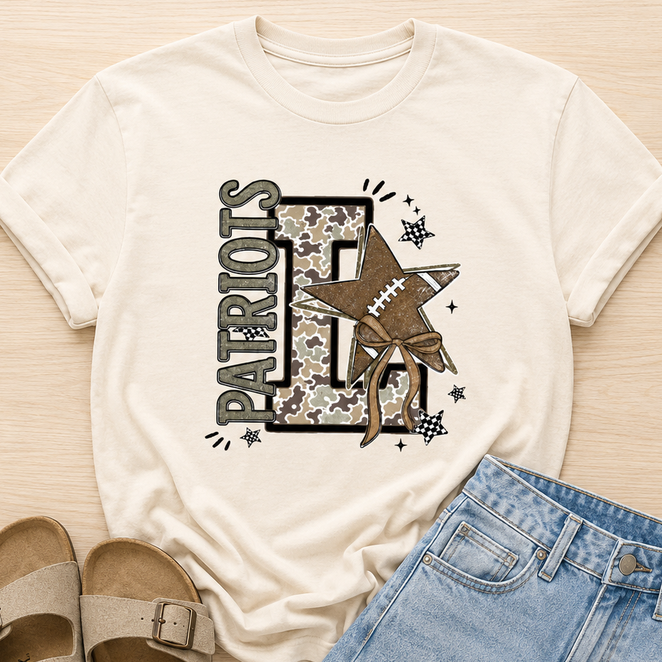 A flat-lay outfit featuring an off-white graphic t-shirt with a 'PATRIOTS' and football star design, styled with light blue denim shorts and beige double-strap sandals on a light wood background.