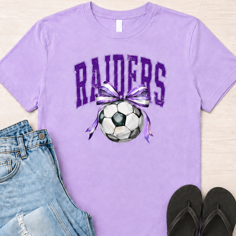 A lavender purple graphic t-shirt featuring the word 'RAIDERS' in purple varsity letters above a soccer ball with a purple and white striped ribbon. The t-shirt is styled in a flat lay with light-wash distressed denim jeans, black flip-flops, and a pair of blue sunglasses on a light wood background.