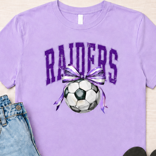A lavender purple graphic t-shirt featuring the word 'RAIDERS' in purple varsity letters above a soccer ball with a purple and white striped ribbon. The t-shirt is styled in a flat lay with light-wash distressed denim jeans, black flip-flops, and a pair of blue sunglasses on a light wood background.