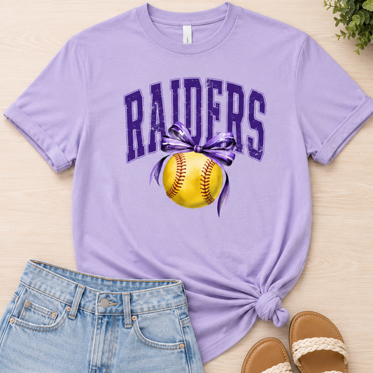A flat-lay outfit featuring a light purple t-shirt with a 'RAIDERS' and softball graphic, styled with light blue denim shorts, brown sandals, and a small green plant on a light wood background.