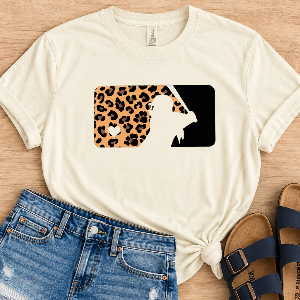 A styled flat-lay featuring an off-white graphic t-shirt with a leopard print and black silhouette design, paired with distressed denim shorts and navy blue slide sandals on a light wood background.