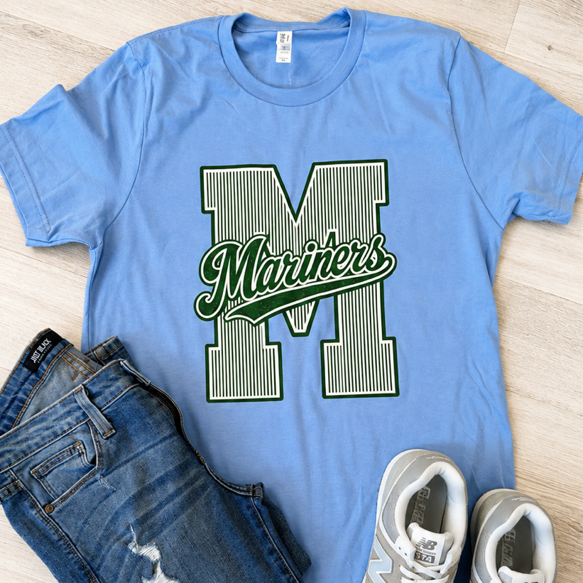 A light blue short-sleeve graphic T-shirt laid flat on a light wood floor, styled with folded blue jeans and white sneakers. The shirt features a large striped letter M with the word 'Mariners' in green script across it.