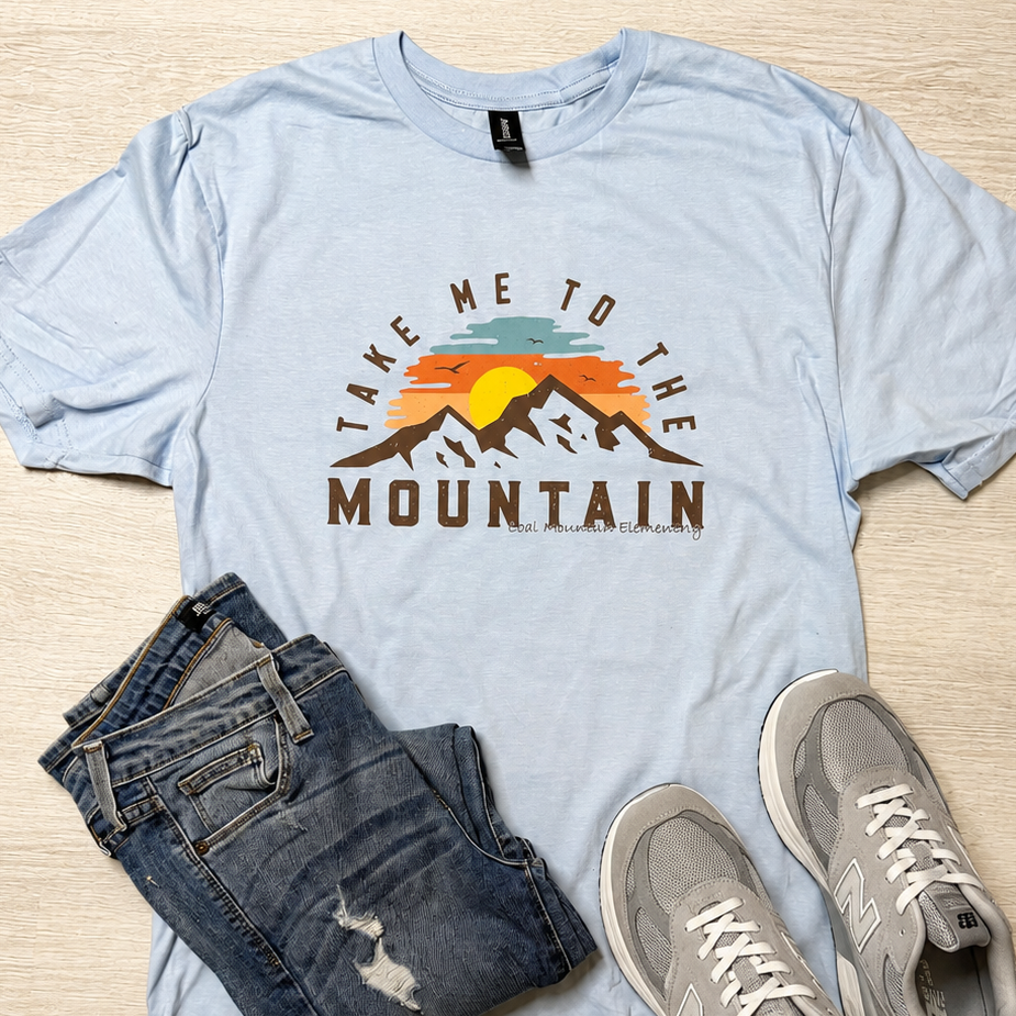 A light blue graphic t-shirt with a 'Take Me to the Mountain' sunset print, styled in a flat lay with dark blue jeans and gray sneakers on a light wood background.