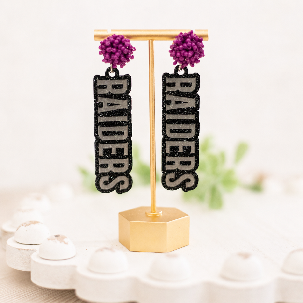 A pair of black and silver glitter drop earrings with purple pom-pom studs, displayed on a gold stand. The vertical dangle charms feature the word 'RAIDERS' in silver glitter letters outlined in black.