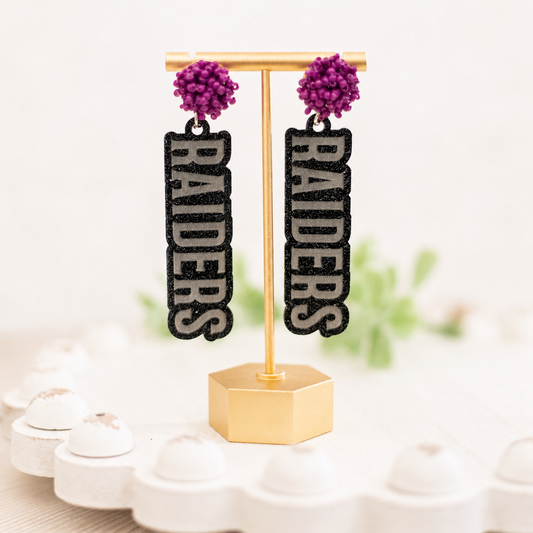 A pair of black and silver glitter drop earrings with purple pom-pom studs, displayed on a gold stand. The vertical dangle charms feature the word 'RAIDERS' in silver glitter letters outlined in black.