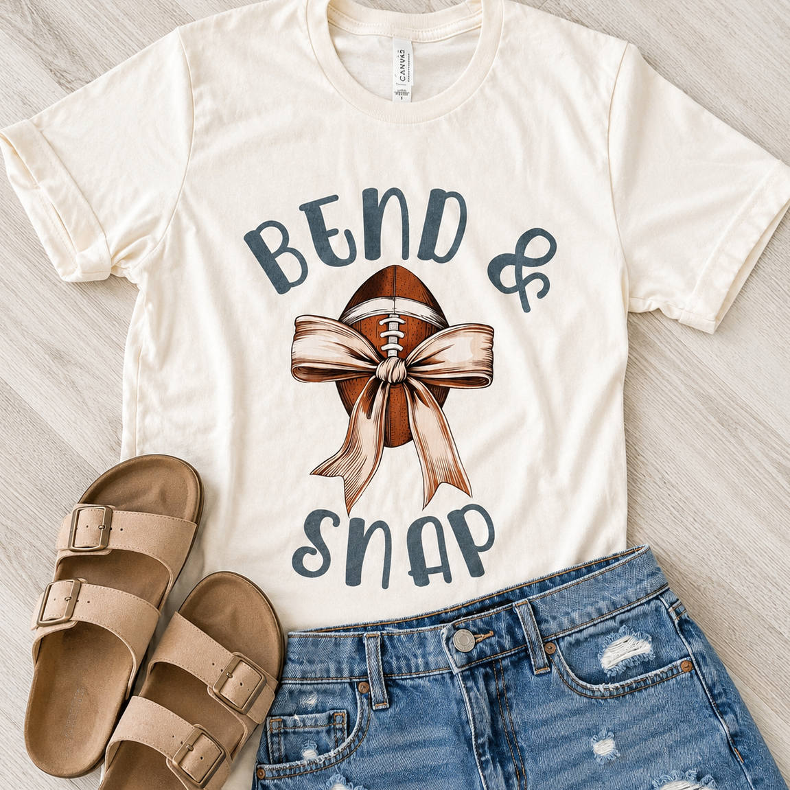 An off-white graphic t-shirt featuring a football with a ribbon and the text 'BEND & SNAP' in navy blue. The t-shirt is styled in a flat lay with beige slide sandals and blue denim shorts on a light wood background.