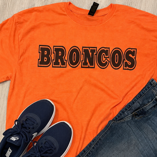 An orange short-sleeve t-shirt with 'BRONCOS' printed across the chest in black outlined lettering, styled in a flat lay with blue jeans and navy blue Nike sneakers on a light wood background.