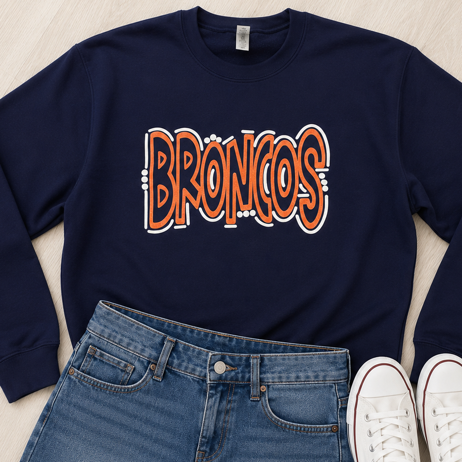 A navy blue crewneck sweatshirt with a stylized orange and white 'BRONCOS' graphic, laid flat on a light wood background alongside blue denim shorts and white low-top sneakers.