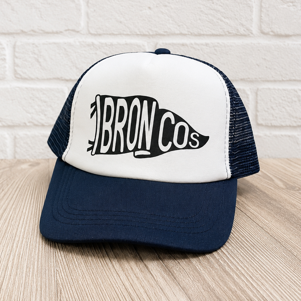 A navy blue and white trucker hat with a black and white 'BRONCOS' graphic on the front, displayed on a light wood surface against a white brick wall background.