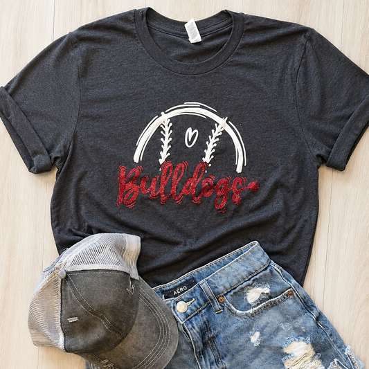 A dark charcoal gray graphic t-shirt featuring a white baseball design and red glittery 'Bulldogs' text, styled in a flat lay with distressed denim shorts and a baseball cap on a light wood background.