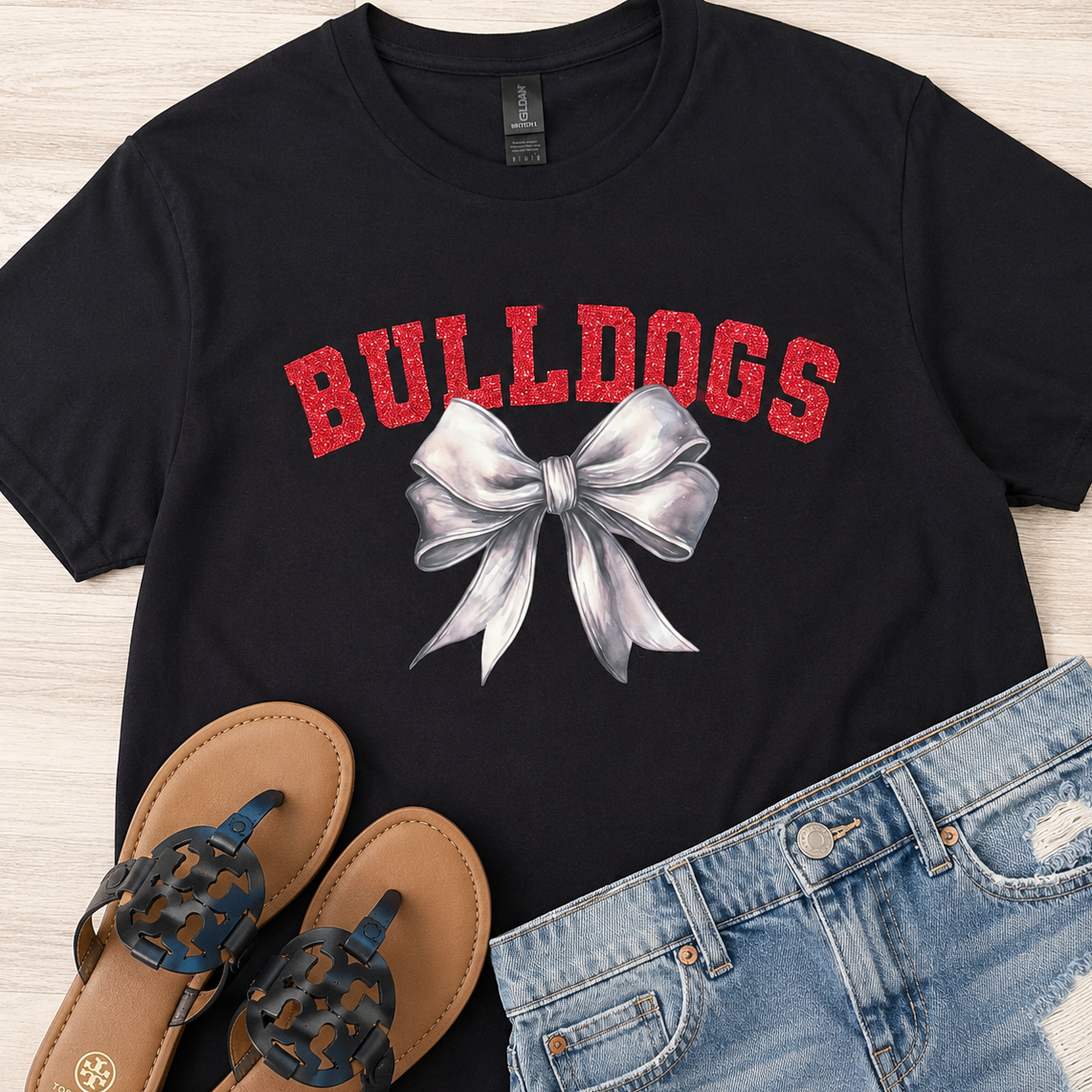 A black short-sleeve graphic T-shirt laid flat on a light wood surface, featuring the word 'BULLDOGS' in red arched letters above a large white bow illustration. Blue denim shorts and brown sandals are styled around it as a flat-lay outfit.