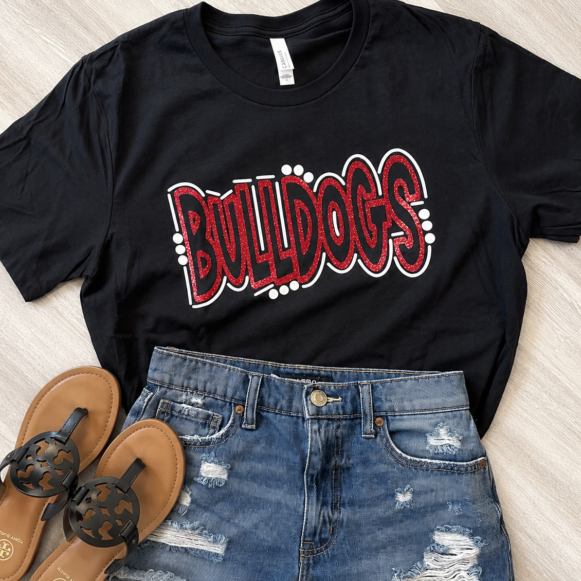A black short-sleeve graphic T-shirt with 'BULLDOGS' in red and white bubble letters, styled in a flat lay with blue distressed denim shorts and brown sandals on a light wood floor.