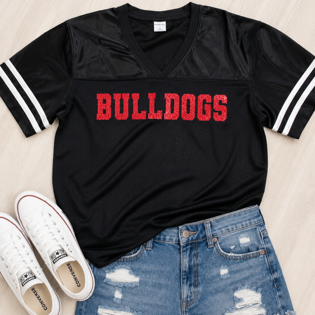 A black short-sleeve V-neck athletic jersey with white stripes on the sleeves and 'BULLDOGS' printed across the chest in red, styled flat-lay with white sneakers and distressed blue denim shorts on a light wood background.