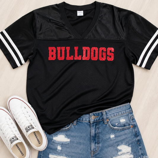 A black short-sleeve V-neck athletic jersey with white stripes on the sleeves and 'BULLDOGS' printed across the chest in red, styled flat-lay with white sneakers and distressed blue denim shorts on a light wood background.