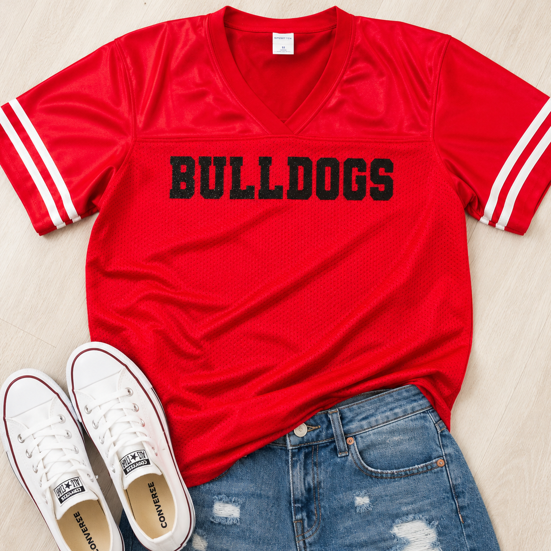 A red short-sleeve V-neck athletic-style shirt with 'BULLDOGS' printed in black across the chest, styled in a flat lay with white distressed denim shorts and white Converse sneakers on a light wood floor.