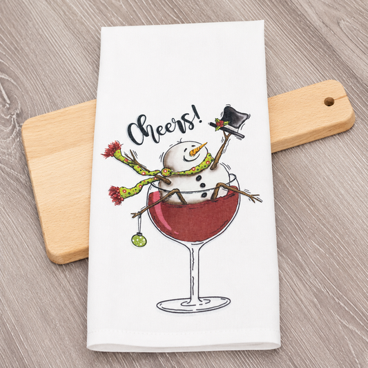 A white kitchen towel featuring a festive illustration of a snowman in a top hat and scarf sitting in a glass of red wine, with the word 'Cheers!' above. The towel is styled on a light wood cutting board over a gray wood-grain tabletop.