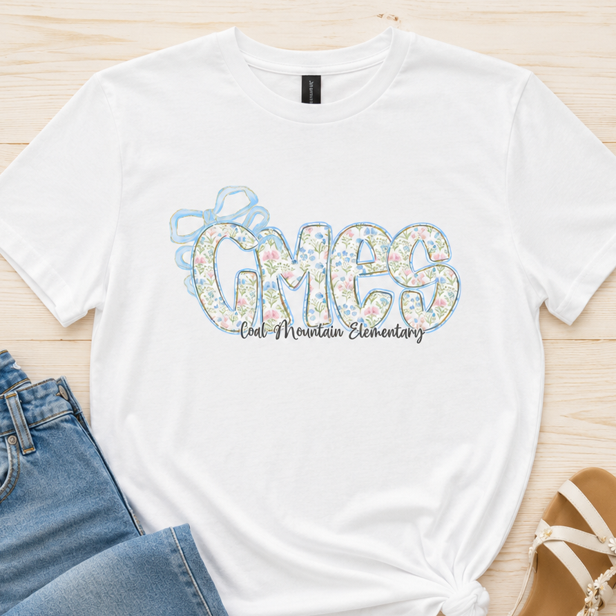 A white graphic t-shirt with 'GMES One Mountain Elementary' printed on the front in a floral pattern, styled in a flat lay with blue jeans and beige sandals on a light wood background.