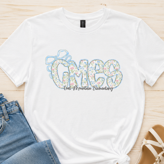 A white graphic t-shirt with 'GMES One Mountain Elementary' printed on the front in a floral pattern, styled in a flat lay with blue jeans and beige sandals on a light wood background.