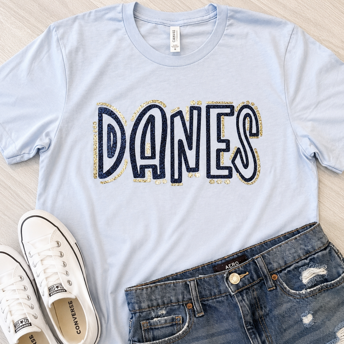 A light blue graphic t-shirt with 'DANES' printed across the chest in navy and gold glitter-style lettering, styled in a flat lay with white Converse sneakers and distressed blue jeans on a light wood background.