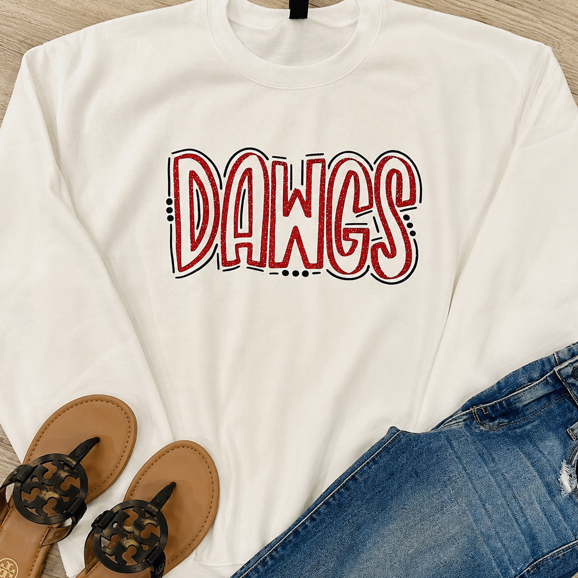 A white long-sleeve sweatshirt with 'DAWGS' printed in red and black bubble letters across the chest, styled in a flat lay with blue jeans and brown thong sandals on a light wood floor.