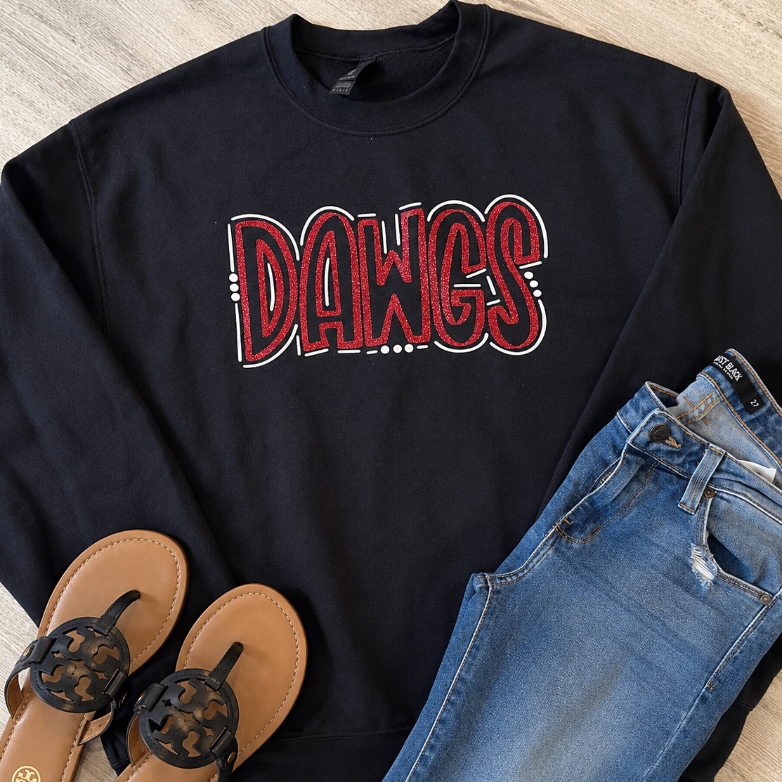 A black crewneck sweatshirt with a red and white 'DAWGS' graphic, styled in a flat lay with blue jeans and brown thong sandals on a light wood floor.