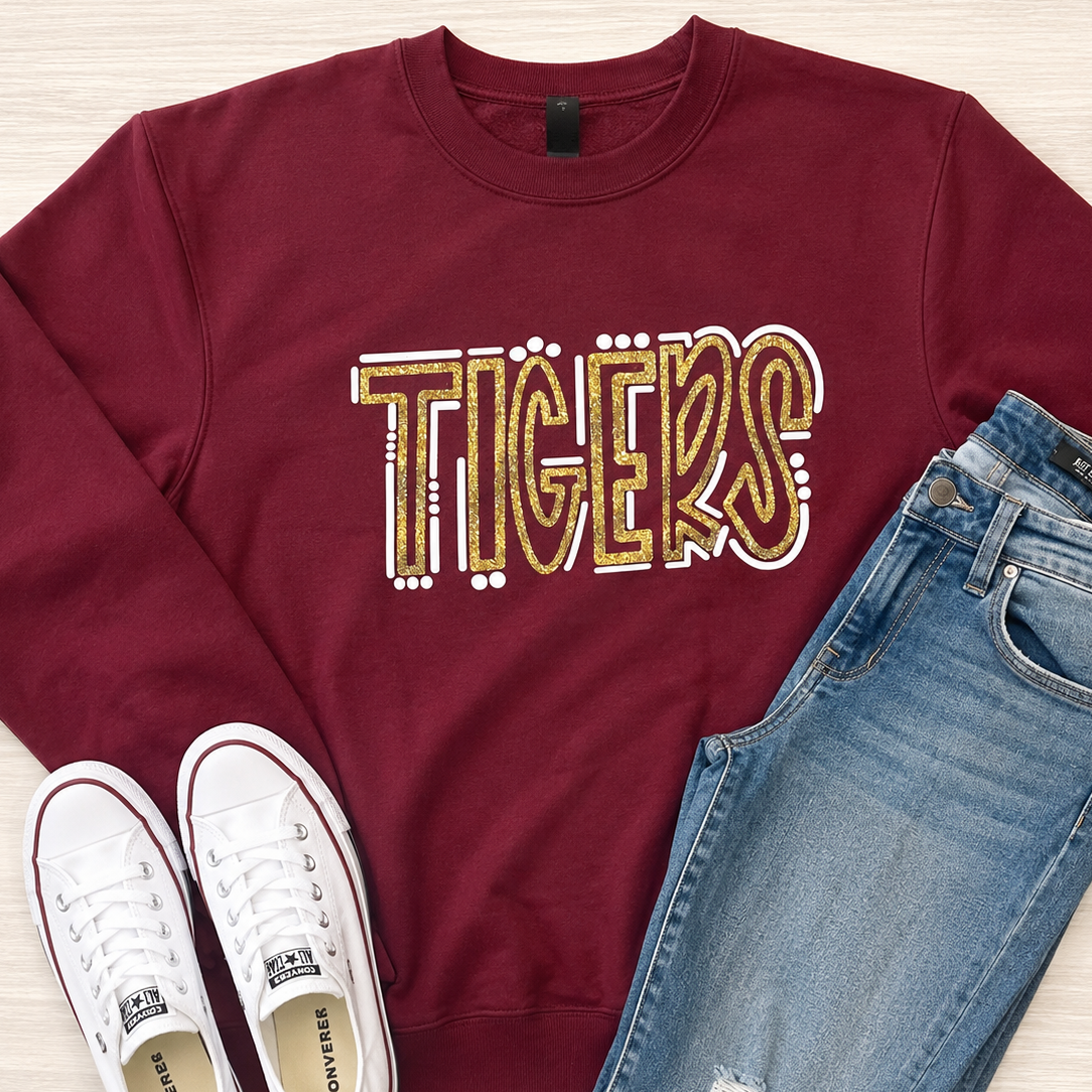 A maroon crewneck sweatshirt with 'TIGERS' printed across the chest in a gold, white, and red patterned font, styled in a flat lay with blue jeans and white Converse sneakers on a light wood background.