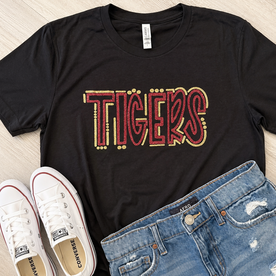 A black short-sleeve graphic T-shirt laid flat on a light wood surface, styled with white Converse sneakers and blue distressed jeans. The shirt has a large 'TIGERS' graphic across the chest in red and gold.