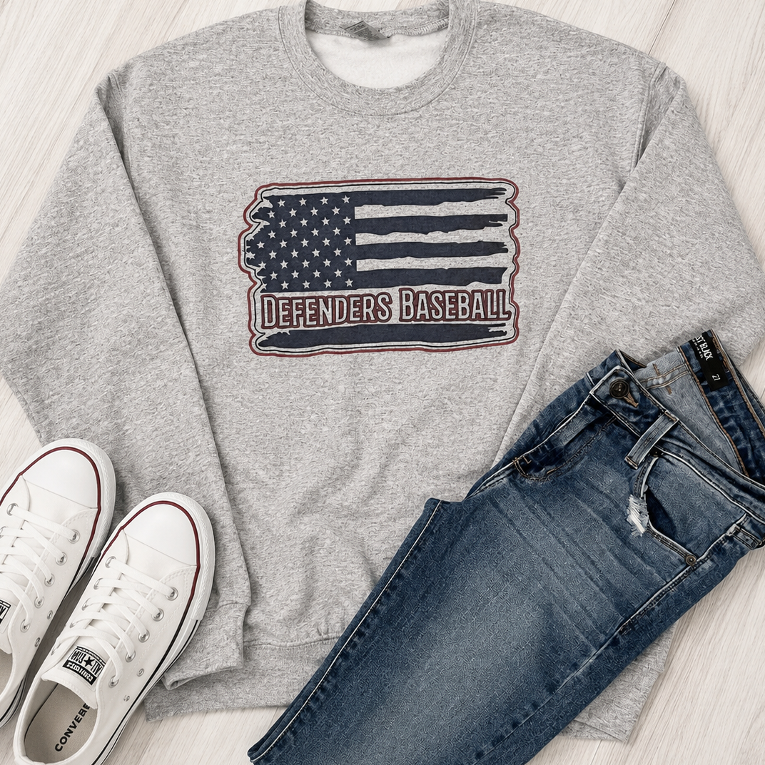 A heather gray crewneck sweatshirt with a distressed American flag graphic and the words 'DEFENDERS BASEBALL' across the chest, styled in a flat lay with blue jeans and white Converse sneakers on a light wood floor.