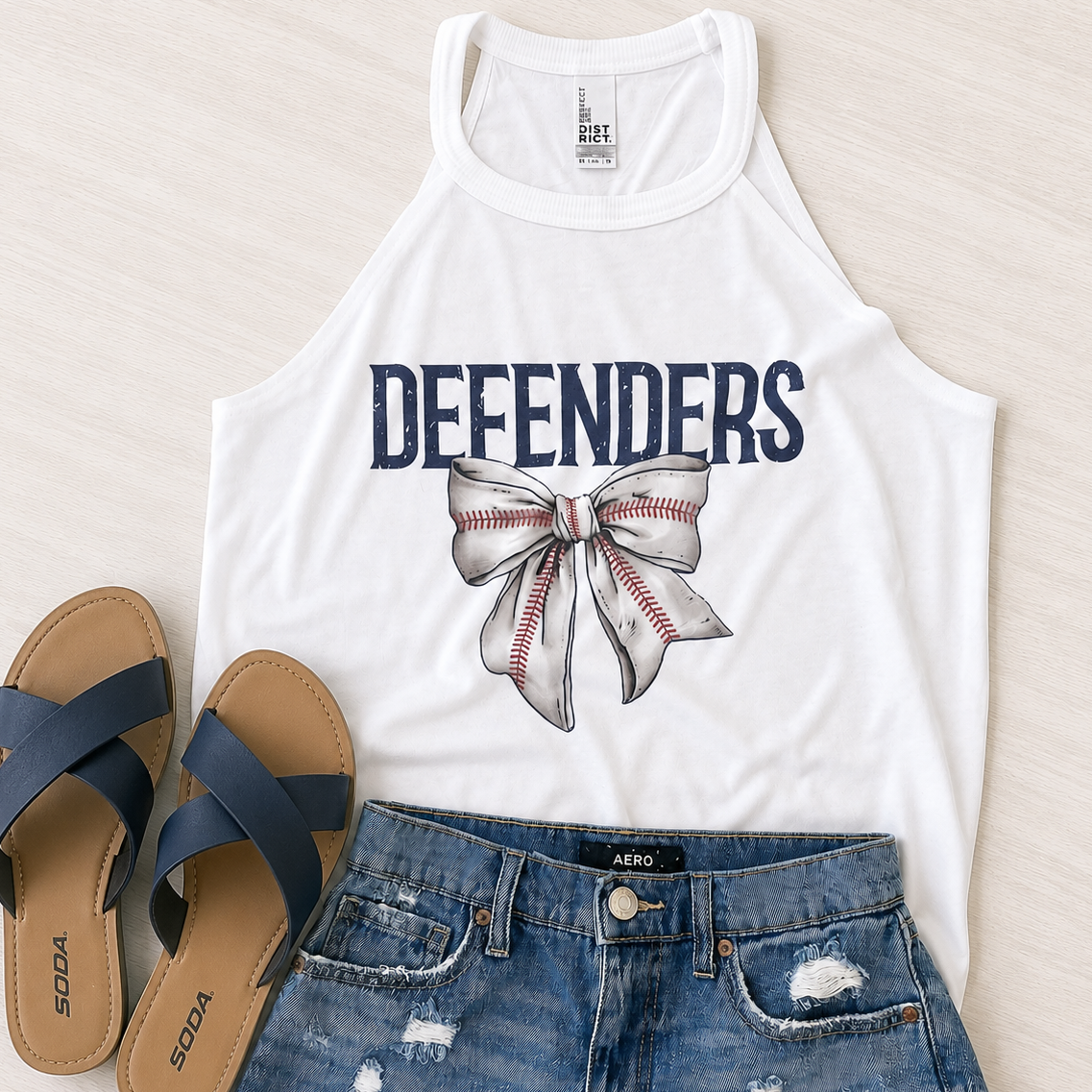 A white sleeveless tank top with 'DEFENDERS' and a baseball-themed bow graphic, styled in a flat lay with blue denim shorts and navy flip-flops on a light wood background.