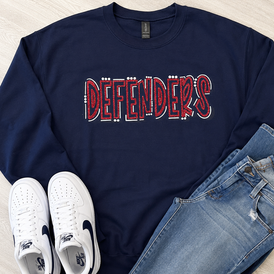 A navy blue crewneck sweatshirt with 'DEFENDERS' printed across the chest in red and white, styled in a flat lay with light blue jeans and white sneakers on a light wood floor.