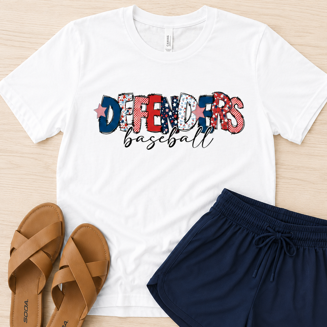 A white short-sleeve graphic T-shirt laid flat on a light wood surface, paired with navy drawstring shorts and tan sandals. The shirt graphic reads "DEFENDERS baseball" with the letters filled in a patriotic red, white, and blue pattern.
