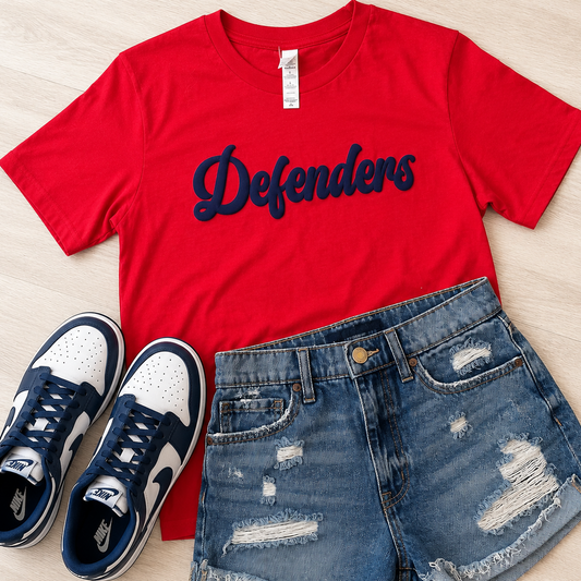 A red short-sleeve T-shirt with 'Defenders' printed in navy across the chest, styled in a flat lay with blue distressed denim shorts and navy-and-white sneakers on a light wood background.
