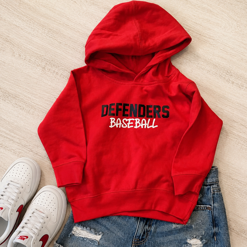 A red pullover hoodie with 'DEFENDERS BASEBALL' printed on the chest, styled in a flat lay with distressed denim shorts and white sneakers on a light wood floor.