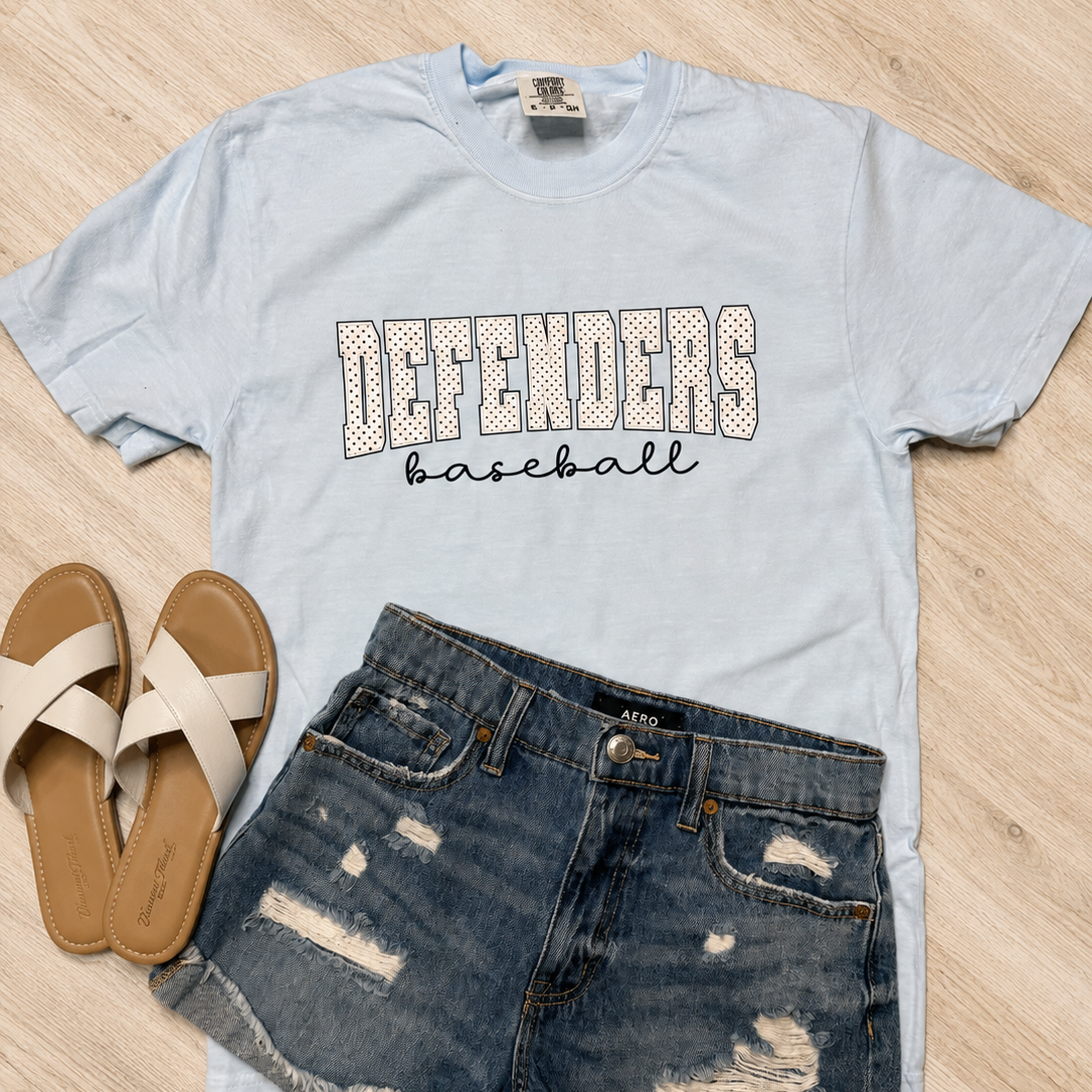 A light blue graphic t-shirt with 'DEFENDERS baseball' printed on the front, styled in a flat lay with distressed denim shorts and beige slide sandals on a light wood floor.