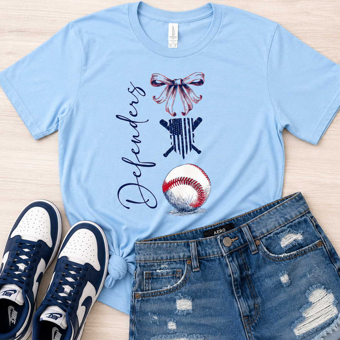 A light blue graphic t-shirt with a baseball-themed design, styled in a flat lay with distressed denim shorts and navy and white sneakers on a light wood background.