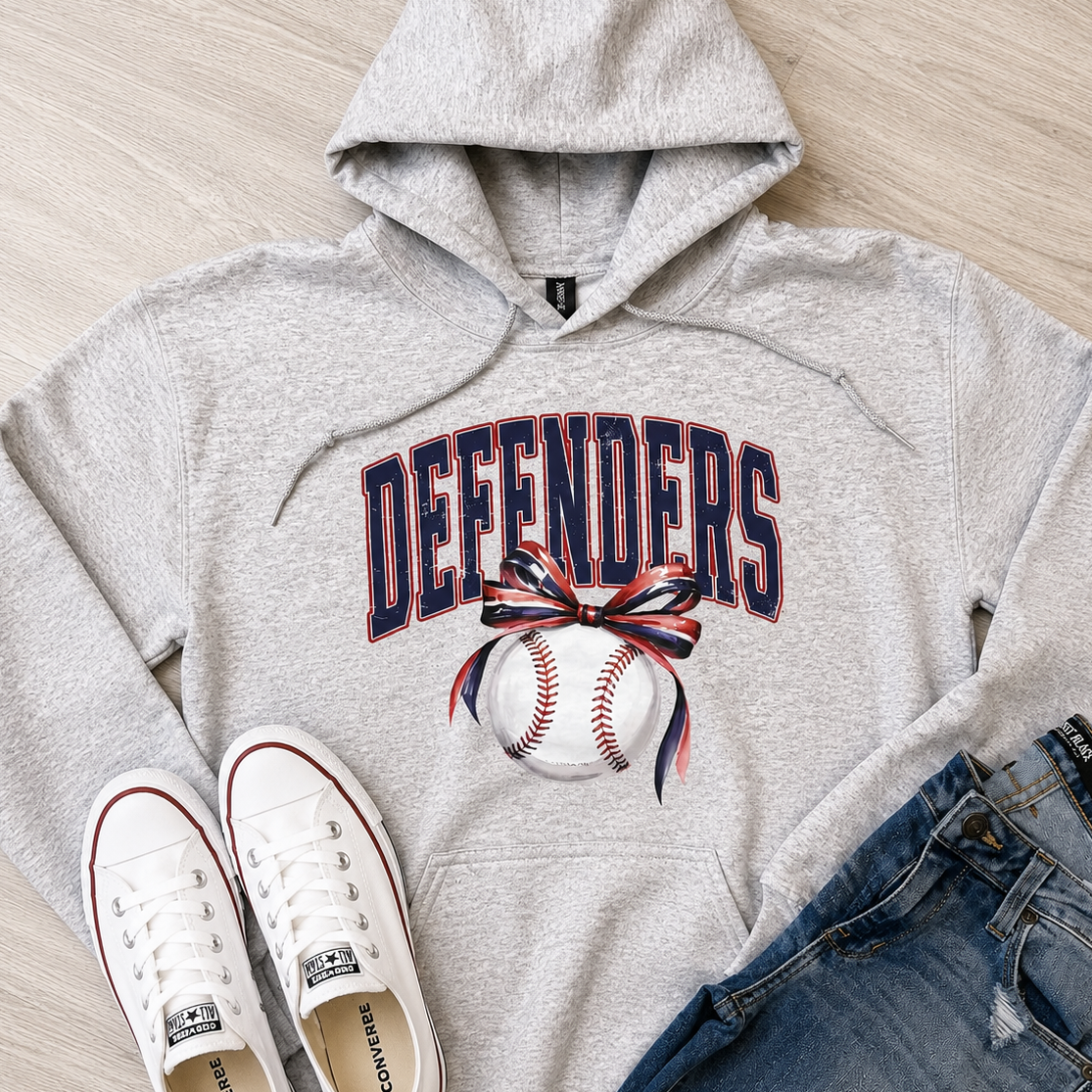 A heather gray pullover hoodie with 'DEFENDERS' and a baseball graphic in navy, red, and white, styled in a flat lay with white Converse sneakers and blue jeans on a light wood floor.