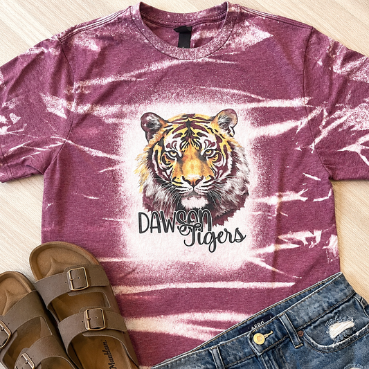 A mauve tie-dye t-shirt with a tiger graphic and the text 'DAWSON Tigers' laid flat on a light wood floor, styled with brown slide sandals and blue jeans.
