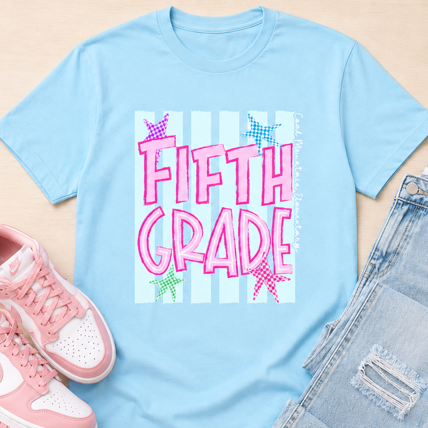 A light blue graphic t-shirt with 'FIFTH GRADE' in pink glitter-style letters, styled in a flat lay with pink sneakers and light-wash distressed jeans on a light wood background.