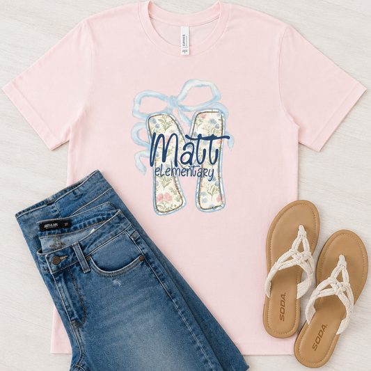 A flat-lay outfit arrangement featuring a light pink short-sleeve t-shirt with a 'Matt Elementary' flip-flop graphic, styled with blue jeans and tan sandals on a light wood background.